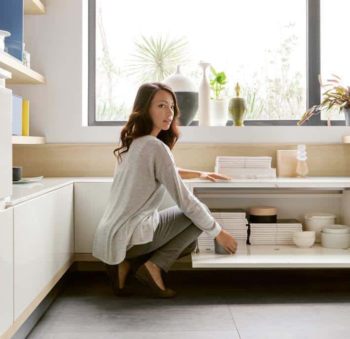 A person in a gray outfit kneels on the floor organizing items on a low shelf in a modern, bright kitchen with a large window.
