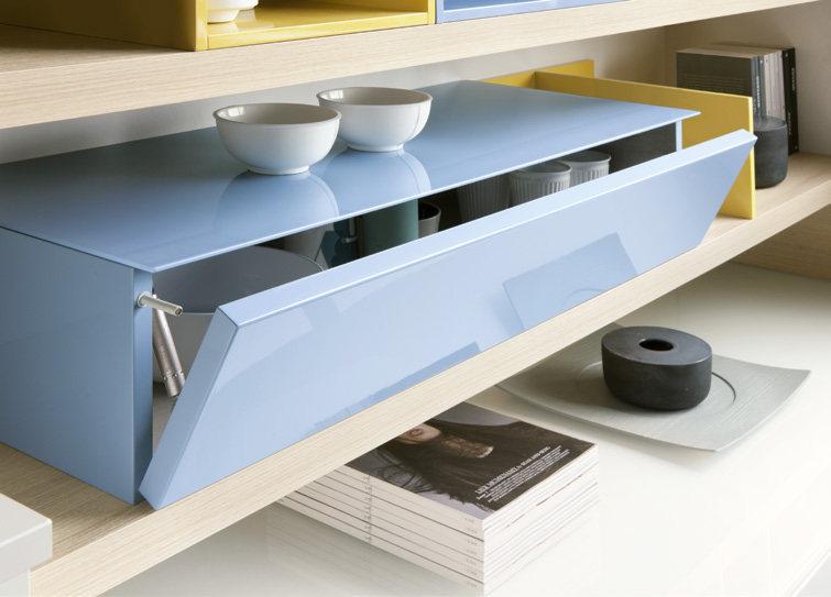 A modern shelving unit with light wood shelves, featuring a glossy blue compartment with a pull-down door, displaying white bowls. Yellow compartments and assorted magazines are also visible.