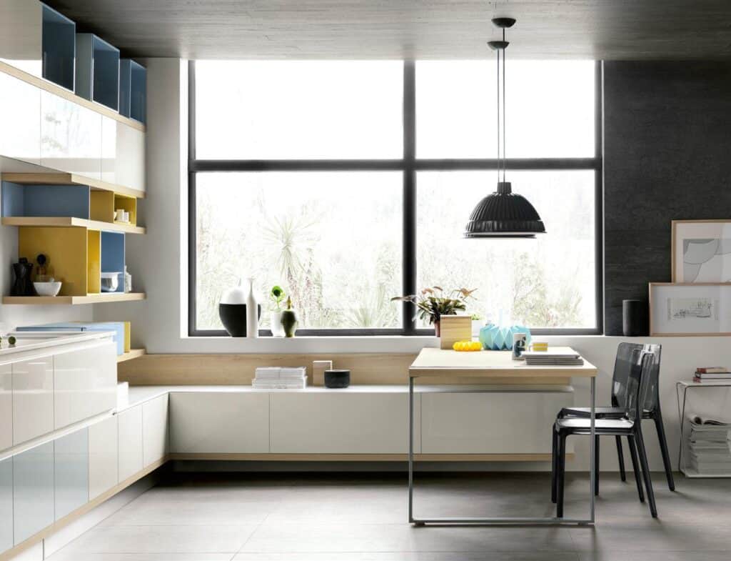 Modern kitchen with large window, sleek white cabinetry, open shelving, a wooden countertop table, two transparent chairs, and a black pendant light.