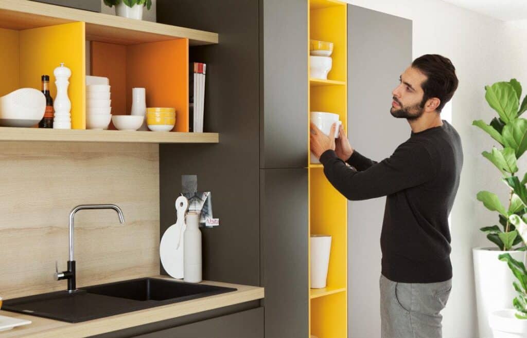 Man organizing dishes in a modern kitchen with open shelves and yellow accents.