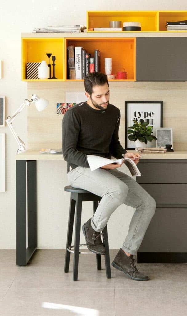 A man sitting on a stool reading a magazine in a modern, minimalist office with yellow and gray shelves, plants, and decor items.
