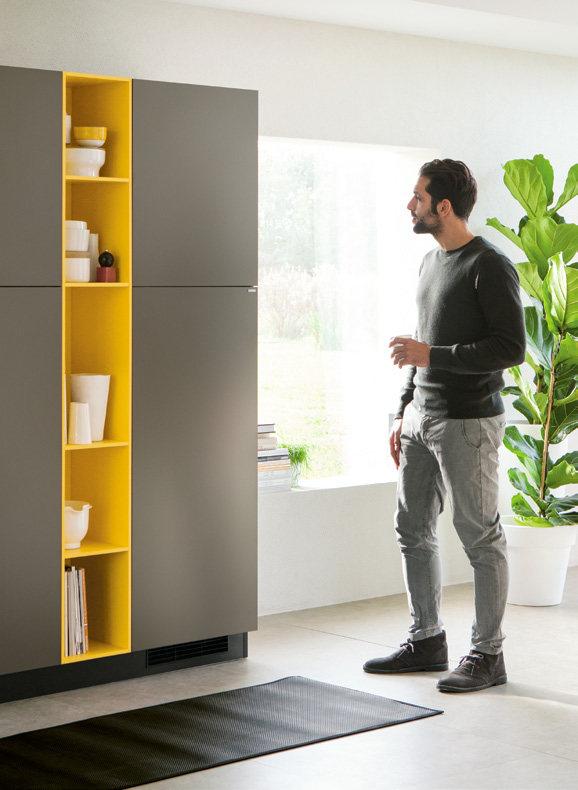 A man in a casual outfit stands near a modern kitchen cabinet with yellow shelves, containing various dishes.