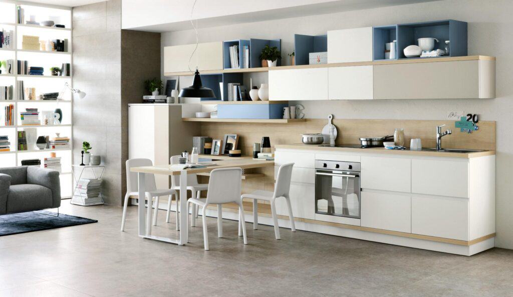 Modern kitchen with light cabinetry, open shelving, a dining table with white chairs, and a built-in oven. Shelves hold decorative items and books, and there is a grey sofa and a lit bookshelf in the background.