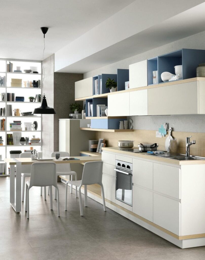 Modern kitchen with sleek white cabinetry, integrated appliances, and a dining area featuring a table with white chairs. Open shelving on the left wall displays books and decor items.