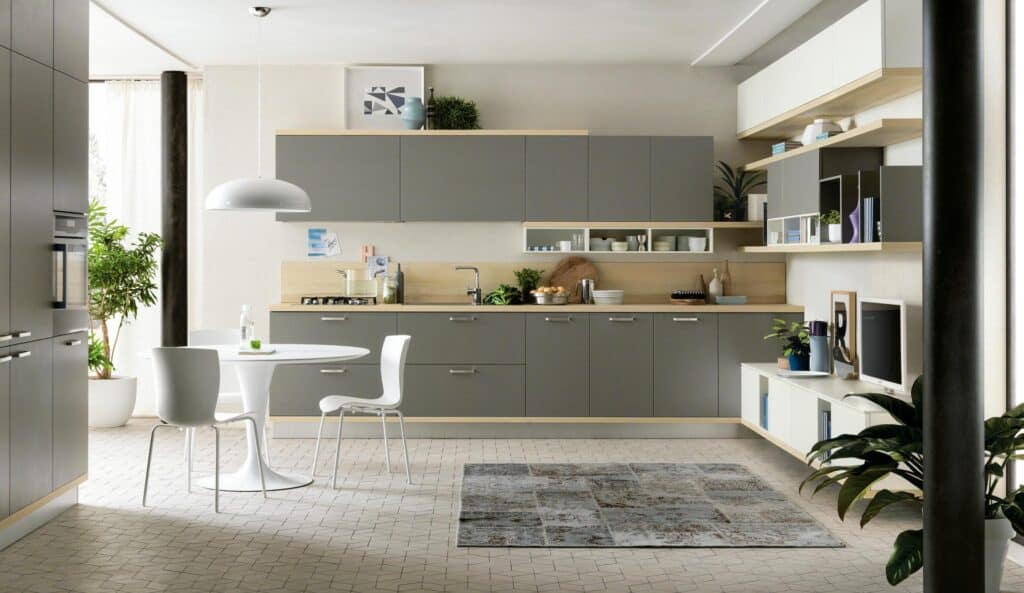 Modern kitchen with gray cabinets, a round white dining table, chairs, plants, and decorative shelves, illuminated by a pendant light.