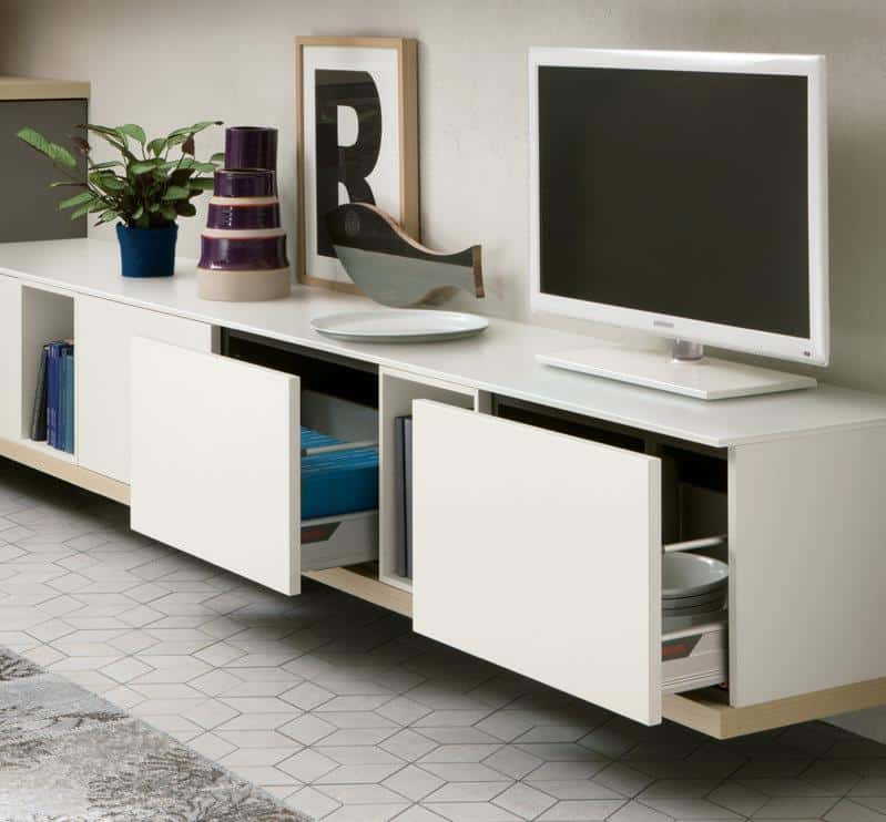 Modern white media unit with open drawers, displaying books and dishes, a TV, decorative items, and a plant on top.