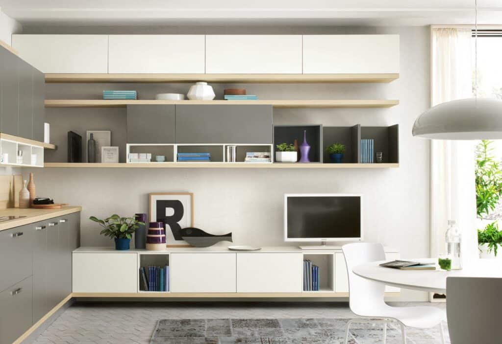 Modern living room interior with grey and white cabinets, wall shelves displaying books and decor items, a flat-screen TV, a round dining table, and bright natural light.