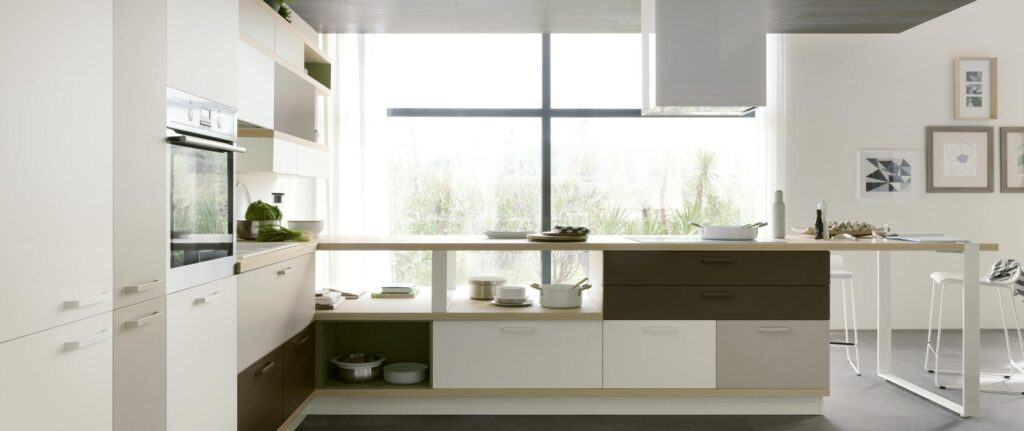 Modern kitchen with light cabinetry, built-in oven, and a large window providing natural light. The kitchen features a central island with open shelving, potted plants, and minimalist decor.