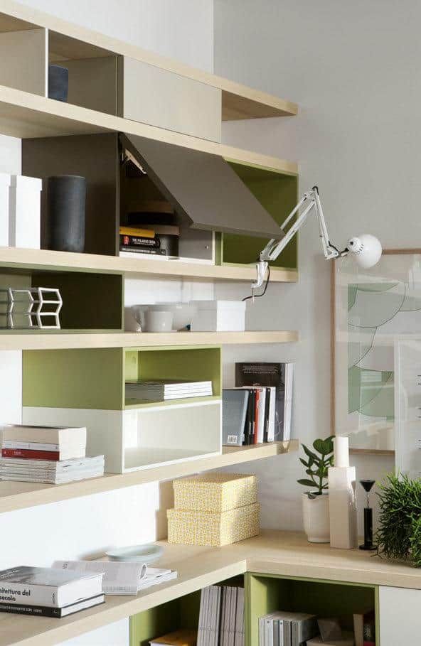 Modern office space with green and beige open shelving, books, decorative items, and a wall-mounted white lamp.