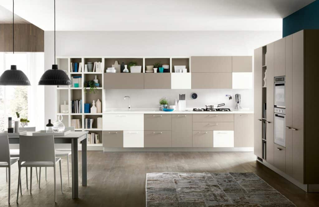 Modern kitchen interior with beige cabinets, white countertops, built-in appliances, open shelving with books and decor, and a dining table with pendant lighting.