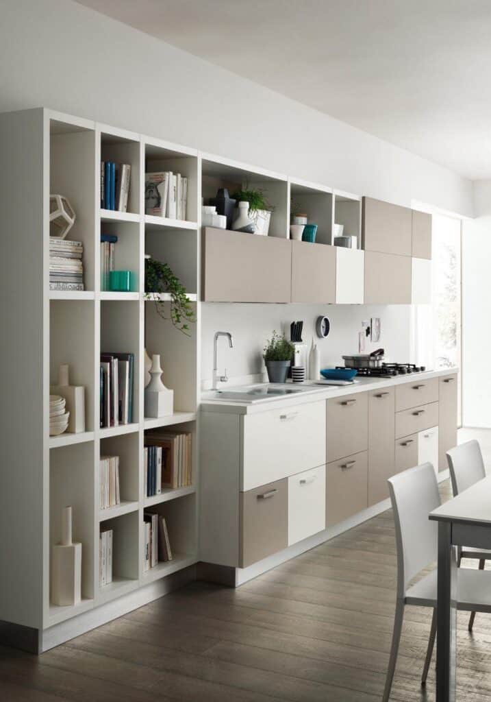 Modern kitchen with beige and white cabinets, a built-in shelving unit displaying books and decor, a sleek countertop with a sink and stove, and a dining table with chairs.