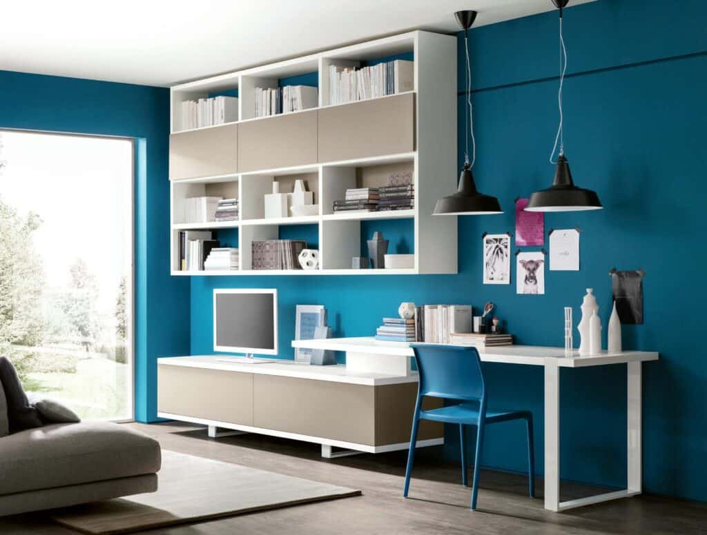 Modern home office with blue walls, white shelving, a desk, a computer, pendant lights, and minimalist decor.