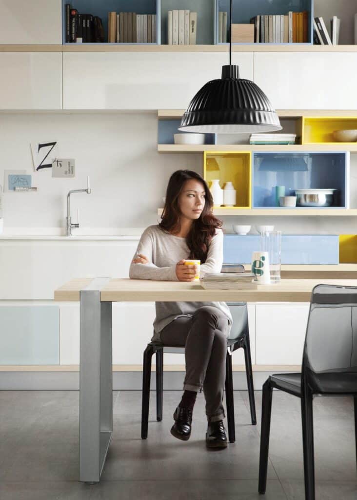Woman sitting at a kitchen table holding a mug, with modern light fixtures and colorful shelves behind her.