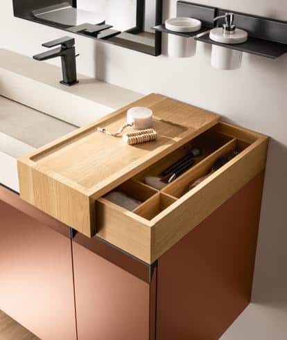 Wooden bathroom drawer partially open, containing grooming items like a brush and razor, near a countertop with a faucet and wall-mounted holders.