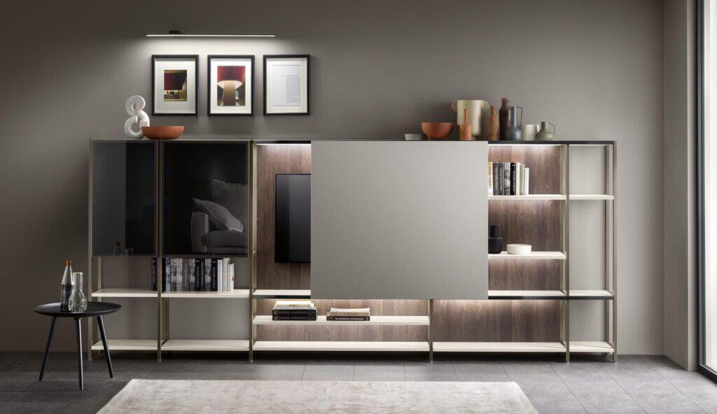 Modern living room with a sleek wall unit featuring open shelves, decorative items, books, and a mounted TV partly covered by a sliding panel, with a small round table displaying a bottle and glass in front.