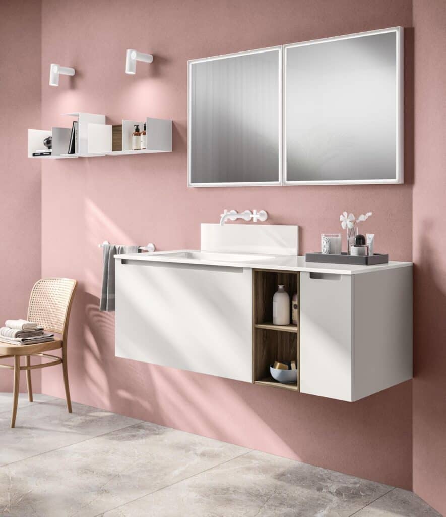 Modern bathroom with a floating white vanity featuring open shelves, set against a pink wall. Large mirror above the sink, wall-mounted lights, and a wicker chair complete the decor.