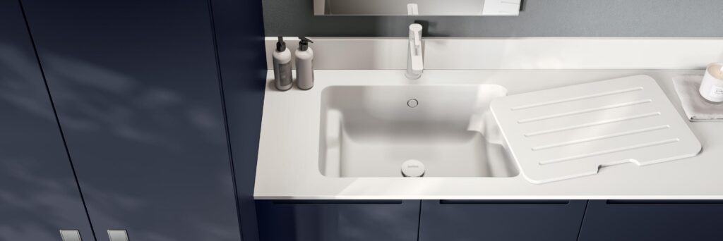 A modern bathroom sink with a white countertop, two soap dispensers, a folded towel, a candle, and a drainer board placed over the sink.