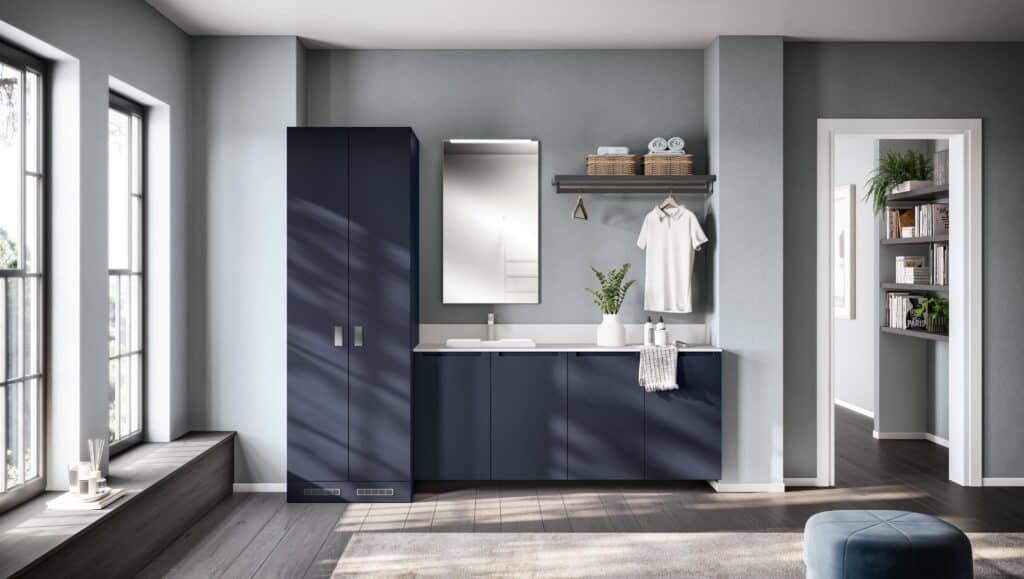Modern laundry room with blue cabinets, a countertop with a sink, and a wall-mounted mirror. A shelf above holds baskets and clothes. A large window with natural light is on the left, and a doorway leads to a room with bookshelves.