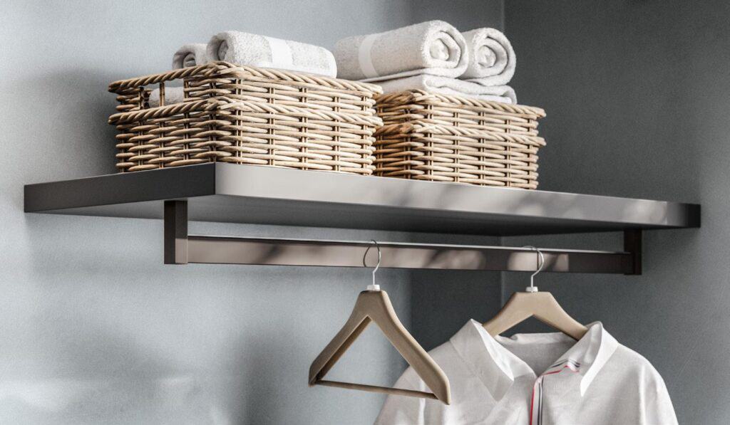 Wall-mounted shelf with two wicker baskets containing rolled towels, a rail beneath holding wooden hangers, one with a white shirt.