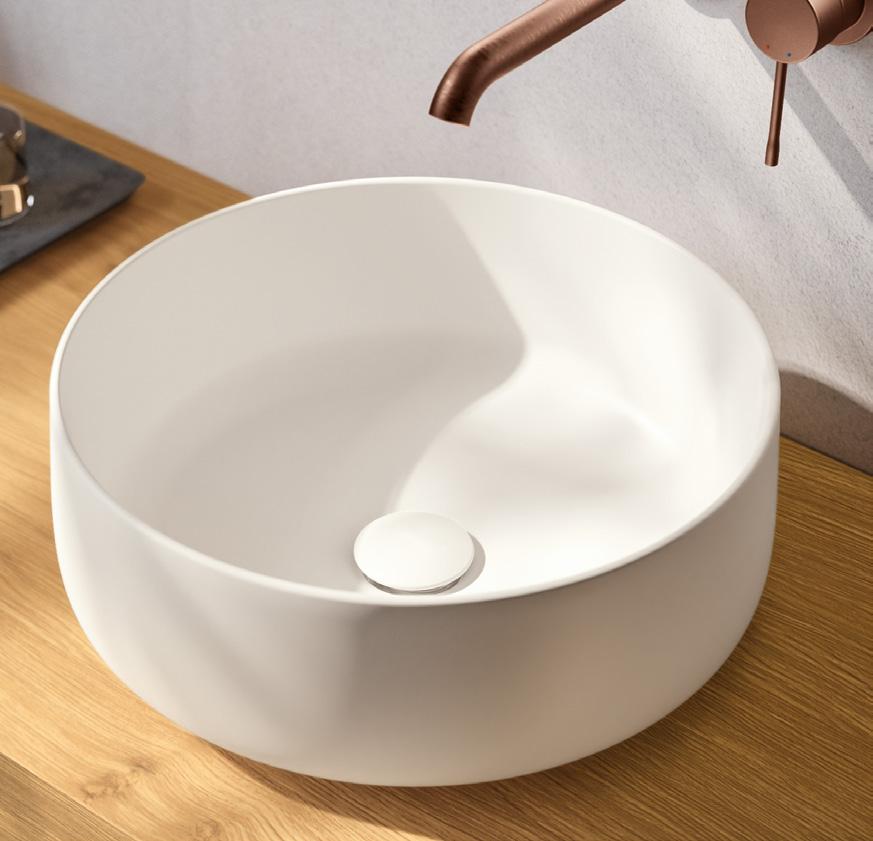 A modern round white ceramic sink on a wooden countertop with a copper faucet mounted on the wall.