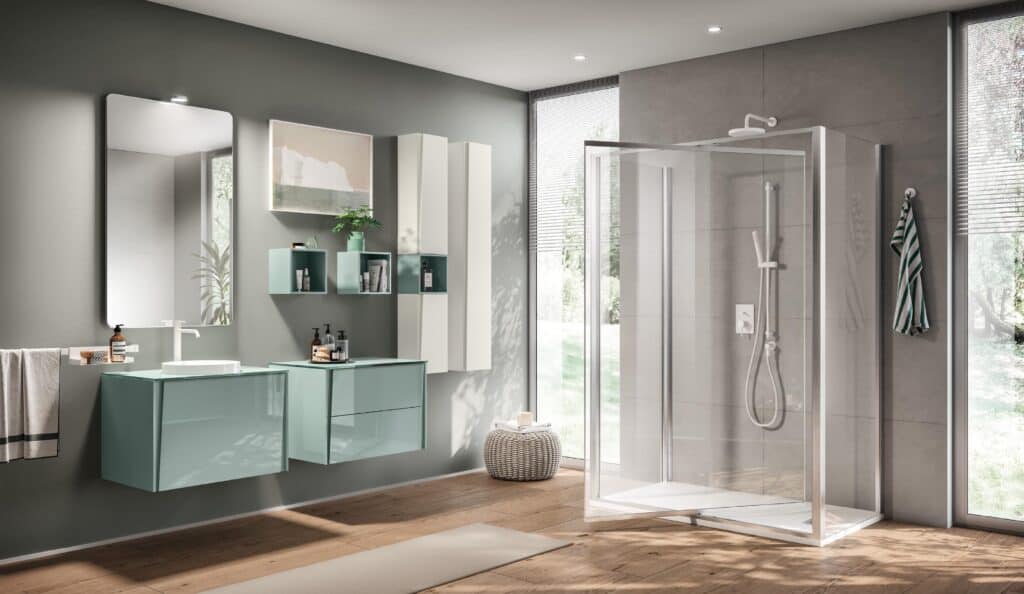 Modern bathroom with a glass shower, teal vanity cabinets, large mirror, wall shelves, potted plant, and wooden flooring.
