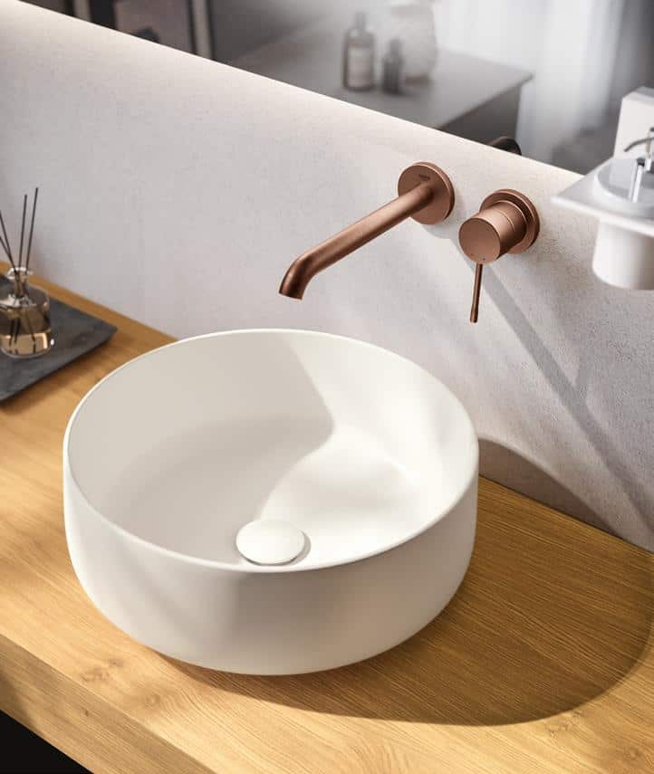 A modern bathroom sink with a round white basin and a wall-mounted copper faucet, set on a wooden countertop.