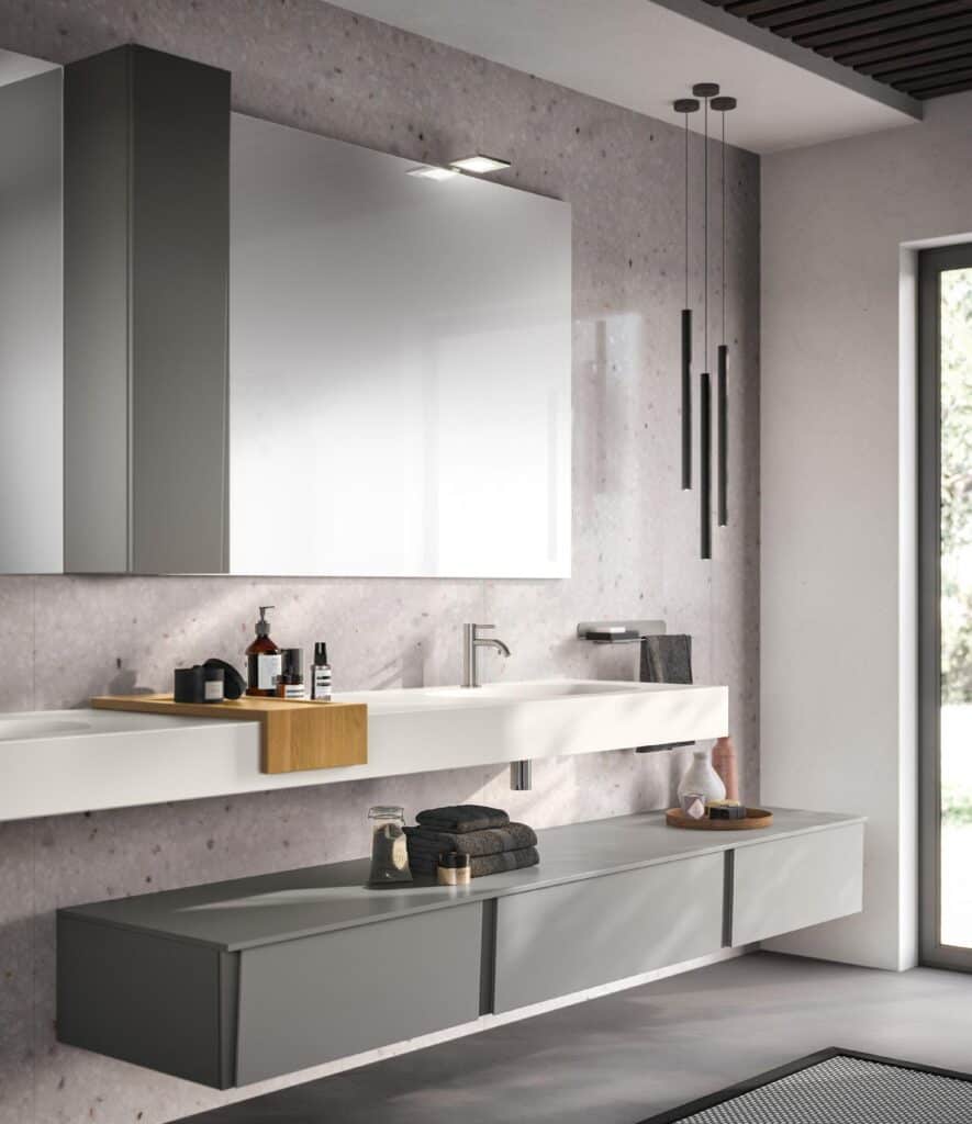 Modern bathroom with a large mirror, sleek white sink, and gray cabinets. Hanging black pendant lights, toiletries, and folded towels add to the minimalist decor.