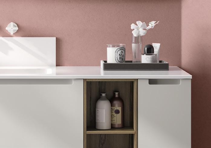 A bathroom counter with white surface, featuring a small open shelf with two bottles, and a tray holding a candle, vase with white flowers, and various toiletries against a pink wall.