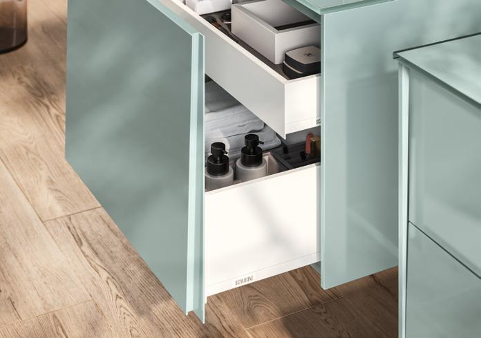 A partially open mint green cabinet drawer revealing organized compartments with bottles and folded items, set against a wooden floor.