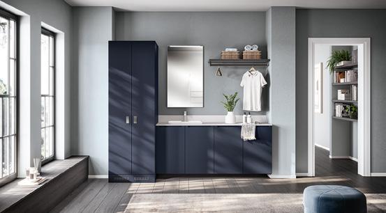 Modern laundry room with dark blue cabinets, a sink, a wall-mounted mirror, and shelves holding laundry items. A white shirt hangs on a hook, and large windows provide natural light.