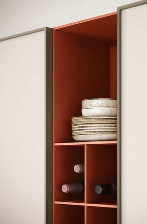 Open kitchen cabinet with orange interior, displaying stacked speckled plates and wine bottles in cubbyholes.