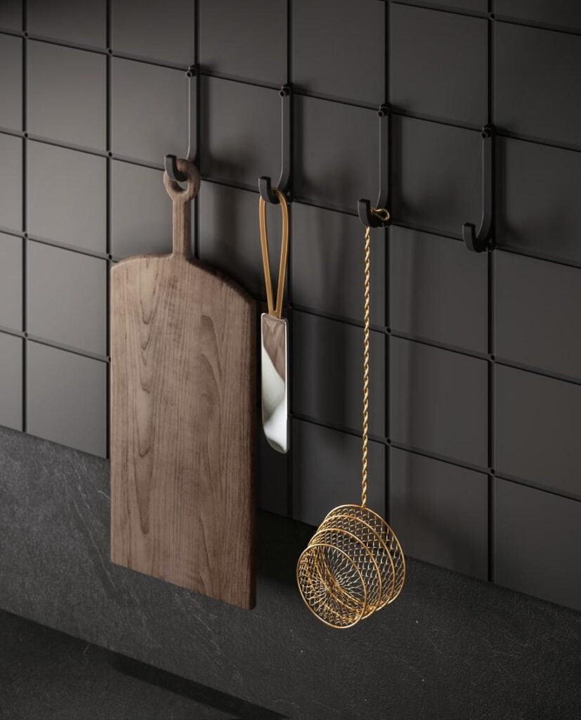 A wooden cutting board, a mirror, and a gold wire basket hang on black hooks against a dark tiled wall.