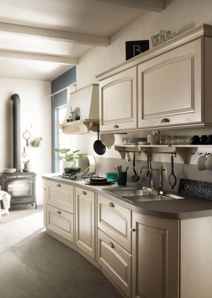 Traditional kitchen with light wood cabinetry, wall-mounted shelves, and utensils hanging above a sink. A stove is next to a decorative exhaust hood, with a small wood-burning stove in the corner.