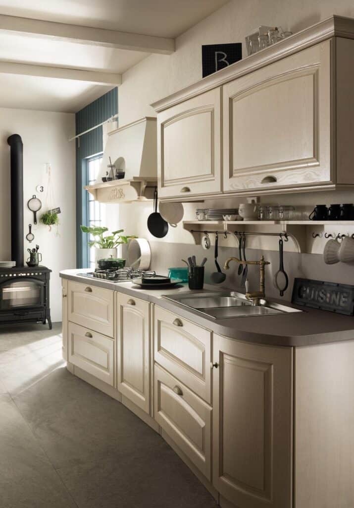 A rustic kitchen with cream-colored cabinetry, a stainless steel sink, a gas stove, and a wood-burning stove.