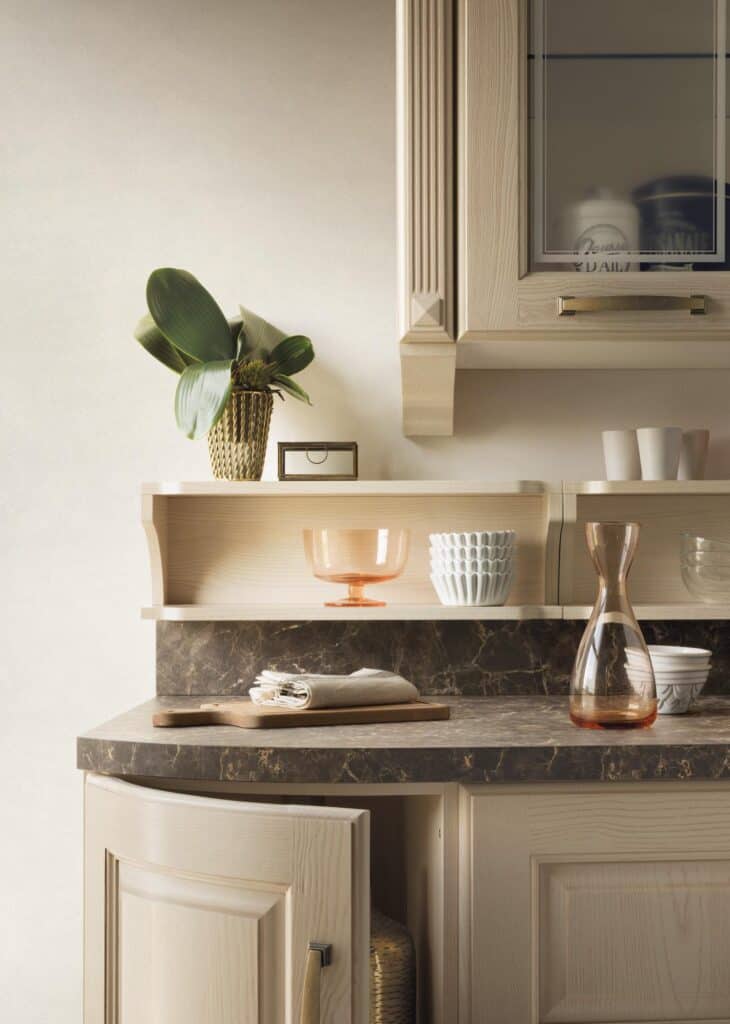 A kitchen countertop with a marble surface, featuring a wooden cutting board with folded cloths, a clear carafe, a decorative plant in a pot, and assorted dishware on shelves.