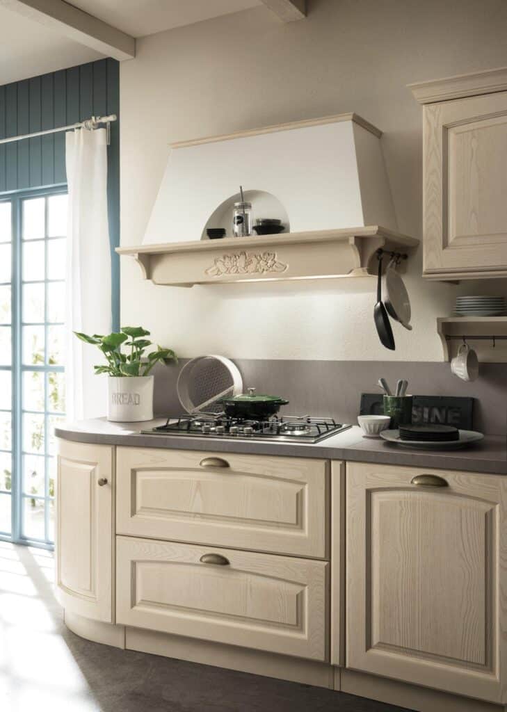 Traditional kitchen with light wood cabinets, a gas stove, white walls, and decorative shelf above the stove. A pot is on the stove, and plants and utensils are displayed on the counter.