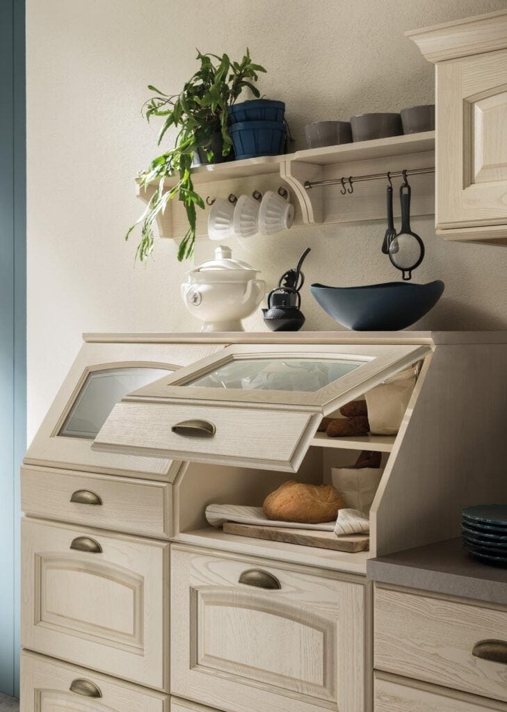 Kitchen with wooden cabinetry, open bread box containing loaves, shelf with potted plants and cups, and countertop with bowls and a tea set.