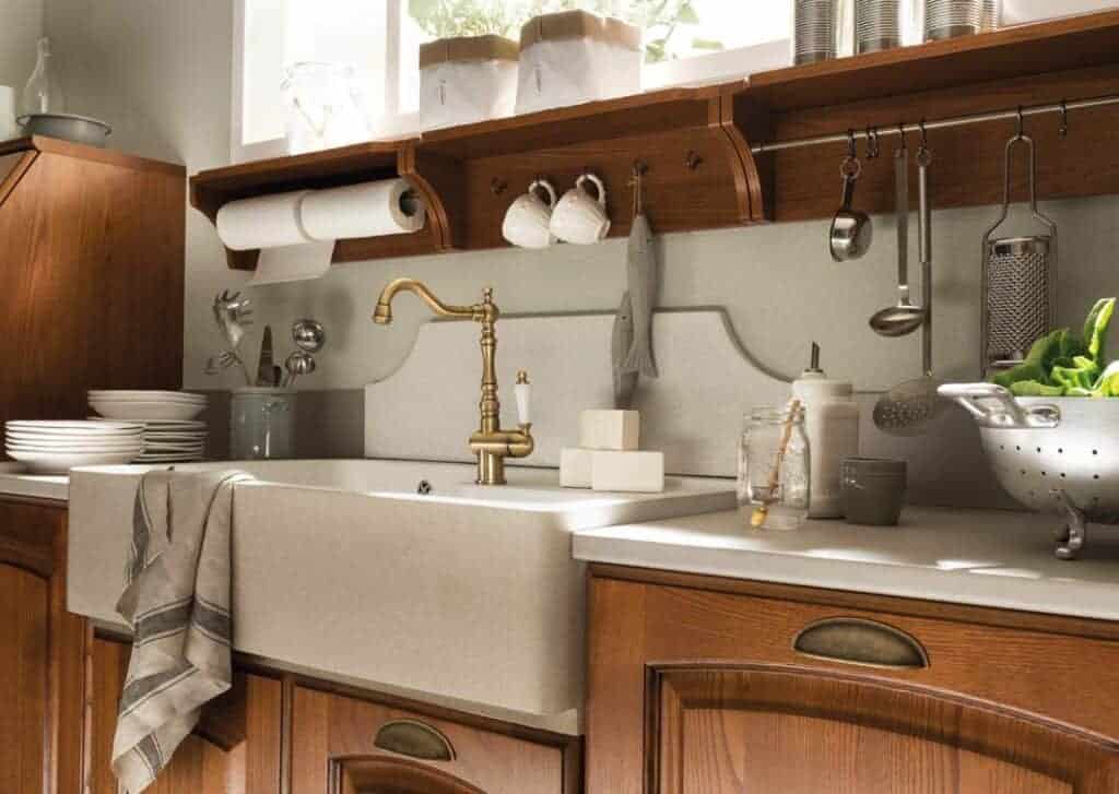 A rustic kitchen sink with a brass faucet, surrounded by wooden cabinets, dishware, and kitchen utensils.