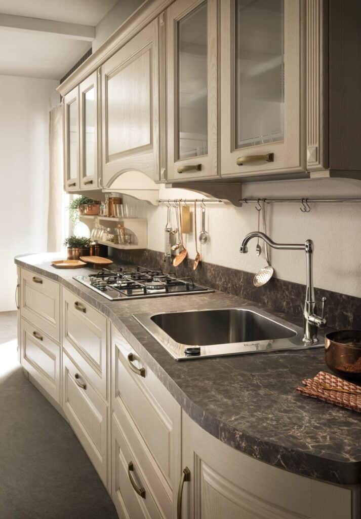 Modern kitchen with beige cabinetry, a marble countertop, stainless steel sink, and gas stove, featuring hanging utensils and copper accents.