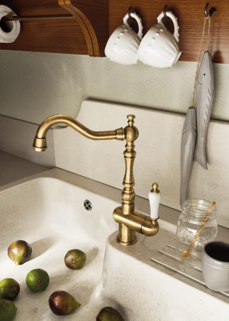 A brass faucet over a cream-colored kitchen sink with figs floating in water. Above, two white mugs hang on hooks and decorative fish hang on the right. A glass jar and spoon are beside the faucet.