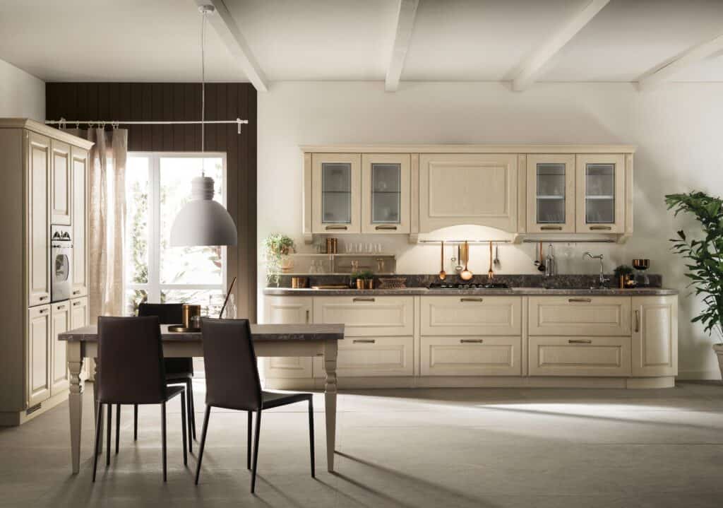 Modern kitchen with light wooden cabinets, dark countertops, and a dining table with two chairs. A large window, overhead lighting, and a potted plant complete the setting.