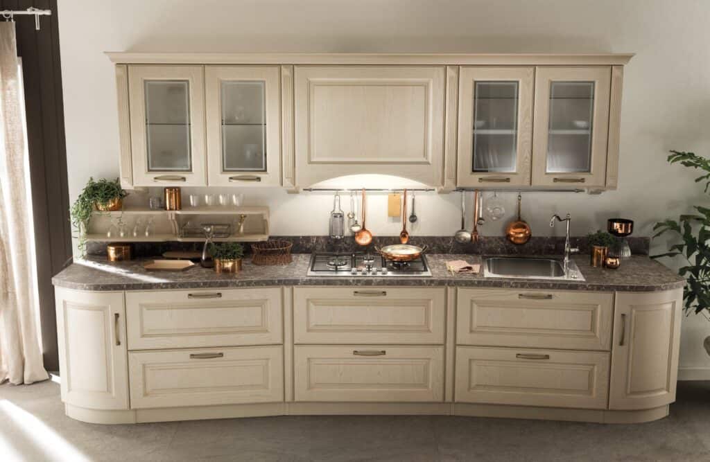 Elegant kitchen with light wood cabinets, glass panel doors, marble countertop, gas stove, sink, copper pots, and plants.