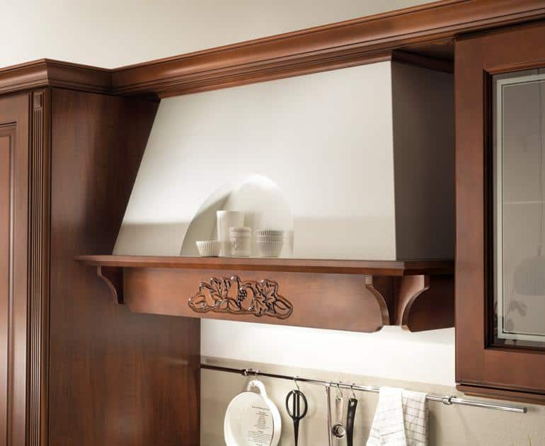 Wooden kitchen range hood with decorative carving and small shelf holding cups.
