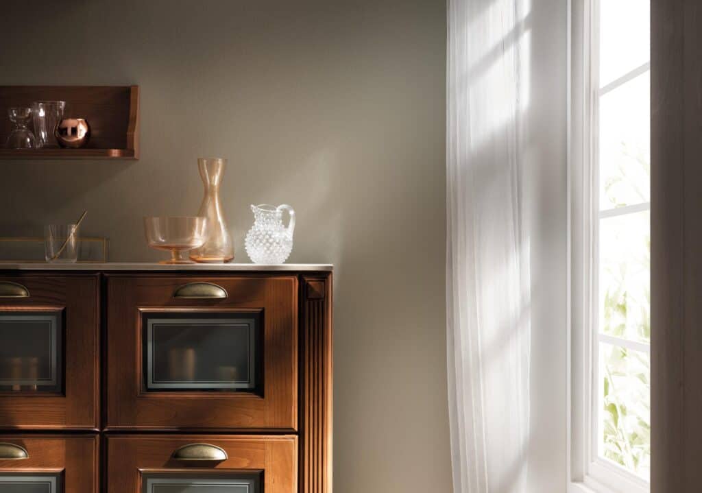 Cabinet with glassware and ceramics, next to a window with sheer curtains.