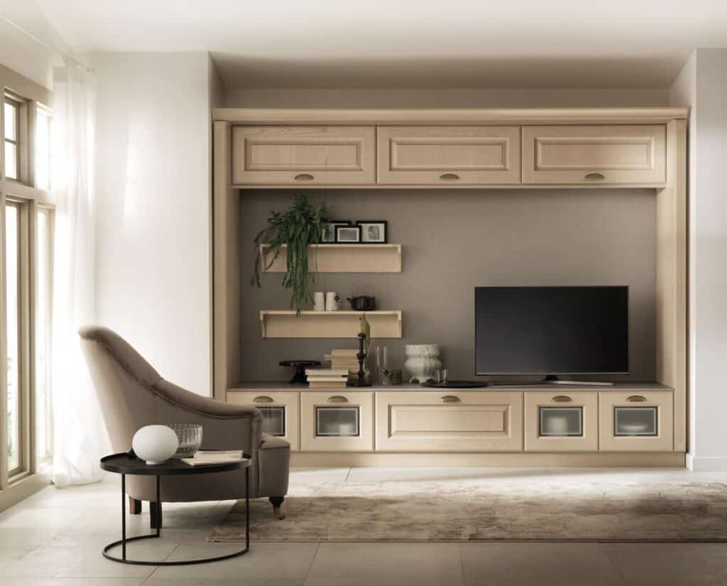 Living room featuring a wall-mounted wooden entertainment unit with a TV, shelves, potted plant, and decorative items, alongside an upholstered armchair and round side table near a window.