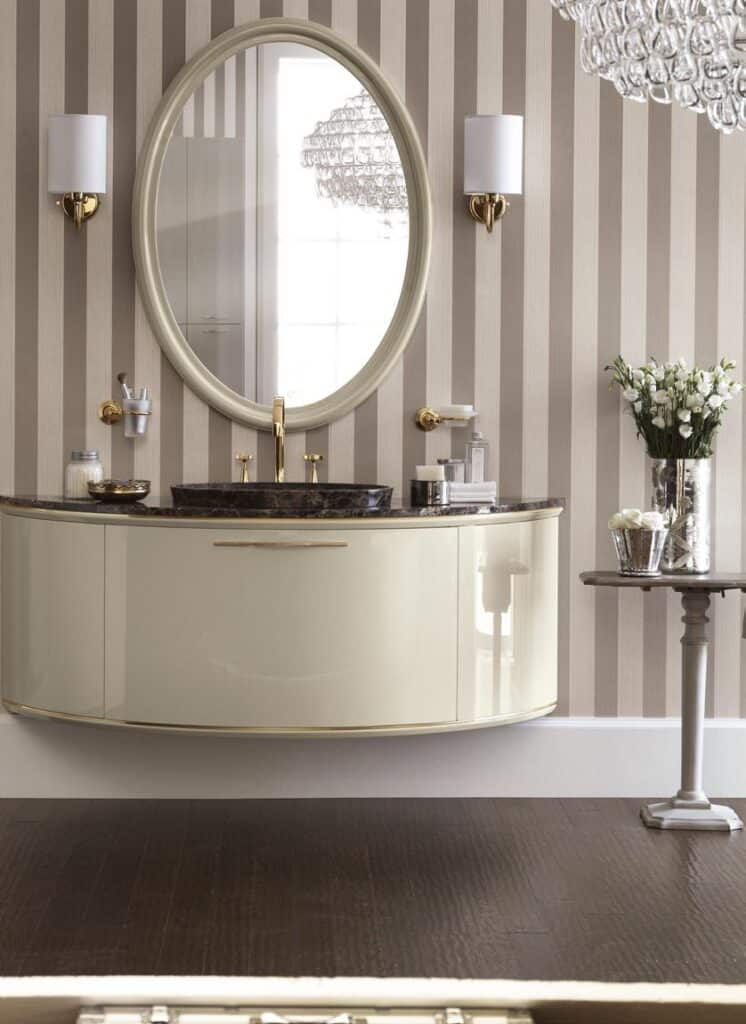 Elegant bathroom with an oval mirror, striped wallpaper, a sleek curved vanity with a marble countertop, and gold fixtures.