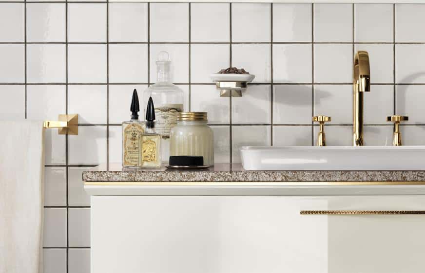 Bathroom sink with gold faucets, glass bottles on the counter, and white tiled backsplash.