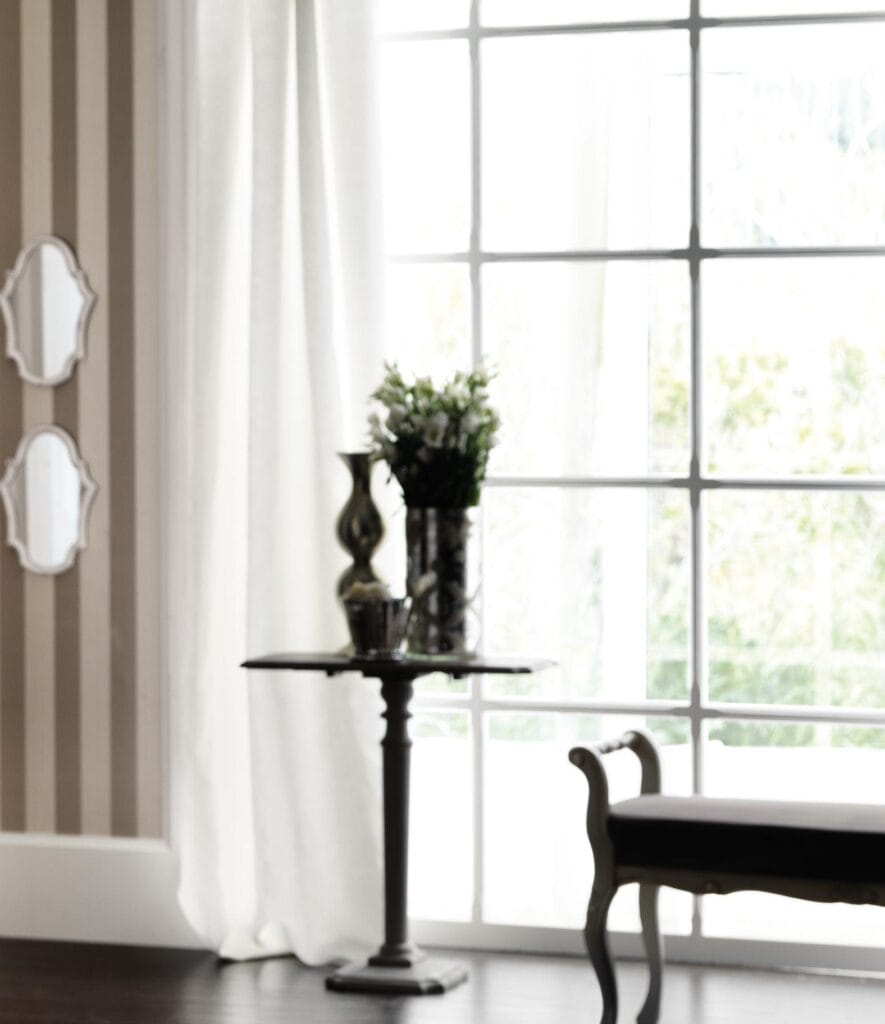 Elegant interior with a small table holding a vase of flowers, a large window with white curtains, and a bench.
