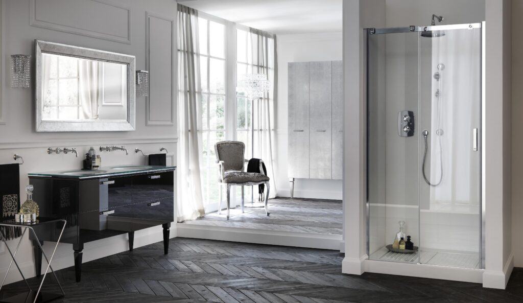 Elegant bathroom with a glass-enclosed shower, black vanity, large mirror, cushioned chair, and pendant light, featuring chevron-patterned wooden flooring and floor-to-ceiling windows with sheer curtains.