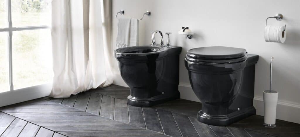 Black bidet and toilet set in a bathroom with a chevron-patterned wood floor, sheer white curtains, and wall-mounted toilet paper holder and towel rack.